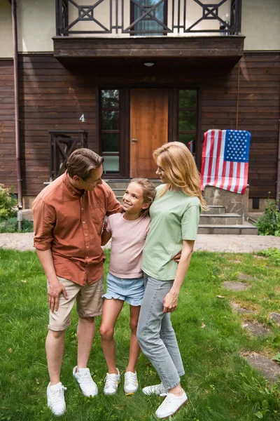 Des parents souriants embrassant leur fille près du drapeau américain flou sur la maison de vacances — Photo de stock