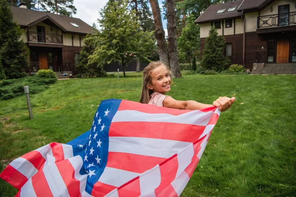 Lächelndes Mädchen blickt in die Kamera, während es mit amerikanischer Flagge im Freien läuft — Stockfoto