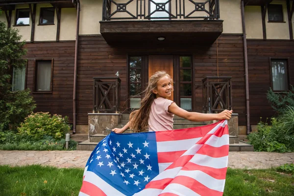 Щаслива дівчина з американським прапором біжить на газоні біля будинку відпочинку — стокове фото