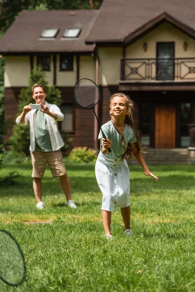 Menina sorrindo jogando badminton perto pai borrado com smartphone no gramado — Fotografia de Stock