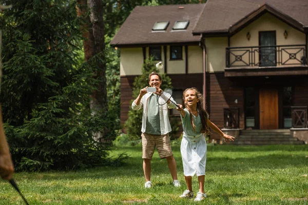 Enfant positif jouant au badminton près du père avec smartphone sur la pelouse — Photo de stock