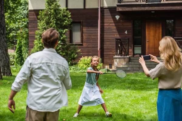 Smiling girl playing badminton near parents with smartphone outdoors — Stock Photo