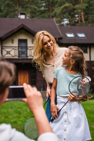 Smiling mother and daughter with badminton rackets standing near blurred father with smartphone — Stock Photo
