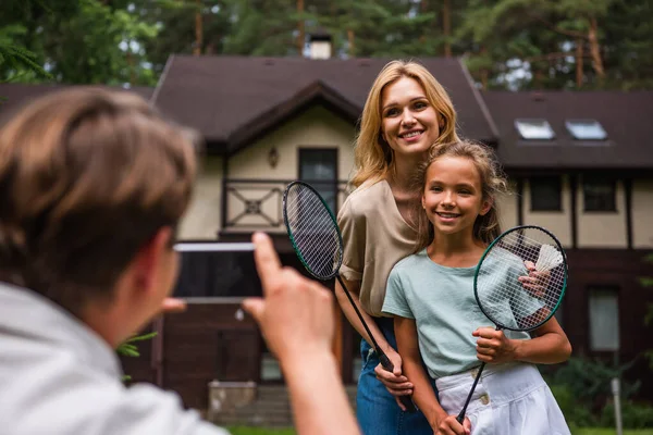 Mother hugging child with badminton racket near husband taking photo on smartphone — Stock Photo