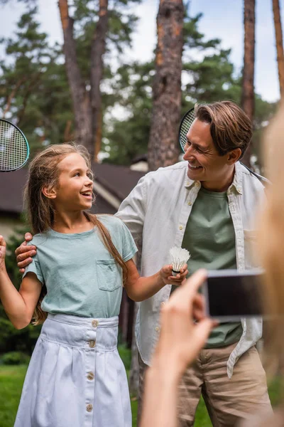 Homme souriant embrasser fille avec raquette de badminton et navette près de femme floue avec téléphone portable — Photo de stock