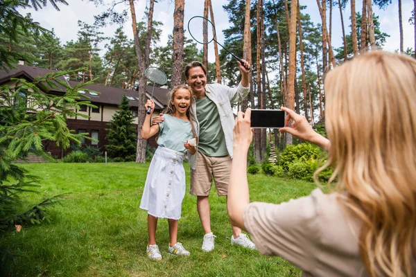 Sonriente padre e hijo sosteniendo raquetas de bádminton cerca de la madre tomando fotos en el teléfono inteligente al aire libre - foto de stock
