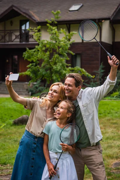 Mulher sorridente tomando selfie no smartphone perto de família alegre com raquetes de badminton — Fotografia de Stock