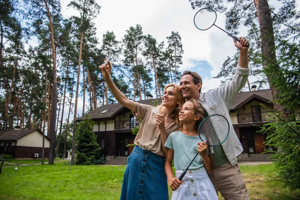 Família sorridente com raquetes de badminton tirando selfie no smartphone durante o fim de semana — Fotografia de Stock