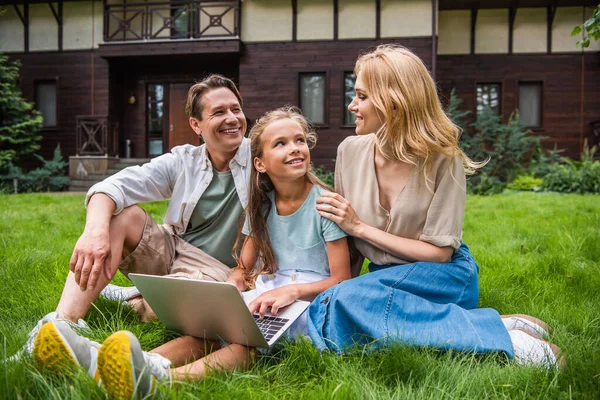 Femme souriante étreignant fille avec ordinateur portable près du mari sur l'herbe — Photo de stock