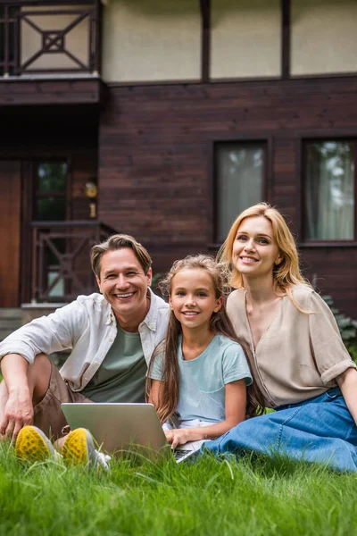 Família sorridente com laptop olhando para a câmera na grama — Fotografia de Stock