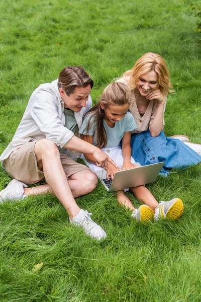 Vista ad alto angolo di uomo che punta con il dito al computer portatile vicino moglie e figlia sull'erba — Foto stock