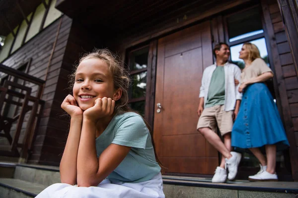 Дівчина дивиться на камеру на сходах поруч з розмитими батьками і будинком відпочинку — стокове фото