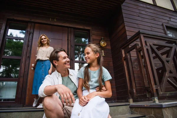Sonriente hombre mirando a la hija cerca borrosa esposa y casa de vacaciones - foto de stock