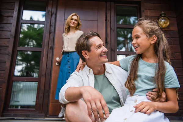 Homme souriant embrassant enfant près de femme floue sur le porche de la maison de vacances — Photo de stock