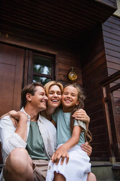 Familia sonriente con los ojos cerrados abrazando en el porche de la casa de vacaciones - foto de stock