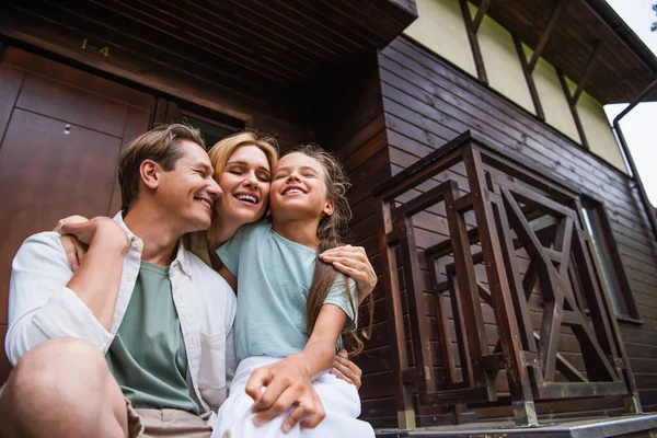 Low angle view of smiling woman with closed eyes hugging daughter and husband near house — Stock Photo