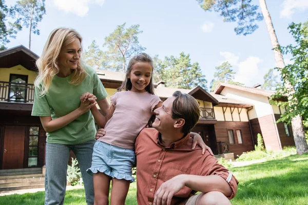 Smiling girl hugging parents on lawn near vacation house — Stock Photo