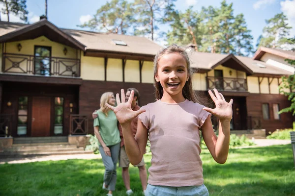 Sonriente chica agitando las manos cerca borrosa padres al aire libre durante las vacaciones - foto de stock