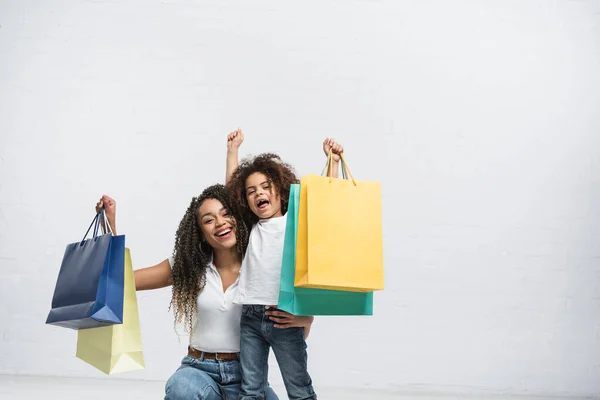 Excited african american mom and daughter with multicolored shopping bags on grey — Stock Photo