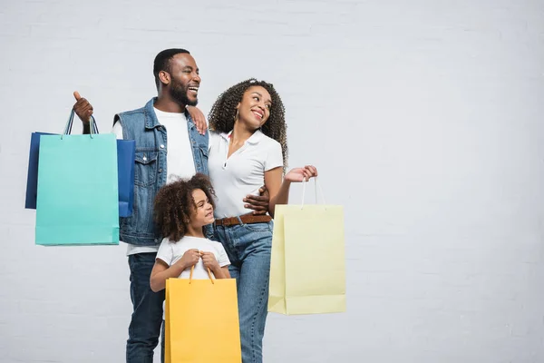 Feliz pareja afroamericana con coloridas bolsas de compras mirando hacia otro lado en gris - foto de stock