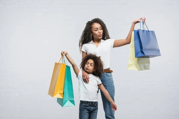 Asombrada mujer afroamericana con hija hinchando las mejillas mientras sostiene bolsas de compras en gris - foto de stock