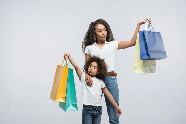 Erstaunt afrikanisch-amerikanische Frau mit Tochter, die Wangen ausbläst, während sie Einkaufstüten auf grau hält — Stockfoto