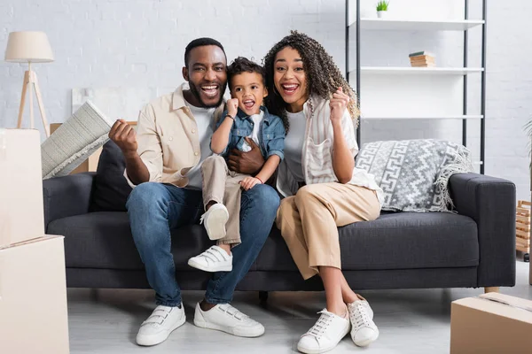 Alegre afroamericano familia mostrando triunfo gesto en sofá en nuevo apartamento - foto de stock