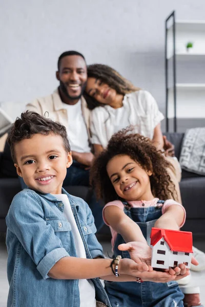 Feliz afro-americanos irmãos mostrando casa modelo perto de pais em fundo turvo — Fotografia de Stock