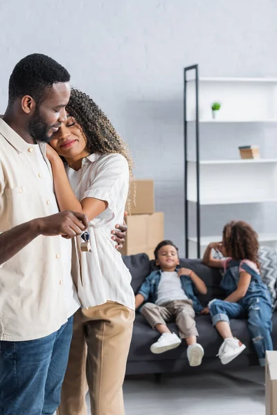 Africano americano uomo con chiave abbracciare felice moglie vicino offuscata bambini in nuovo appartamento — Foto stock