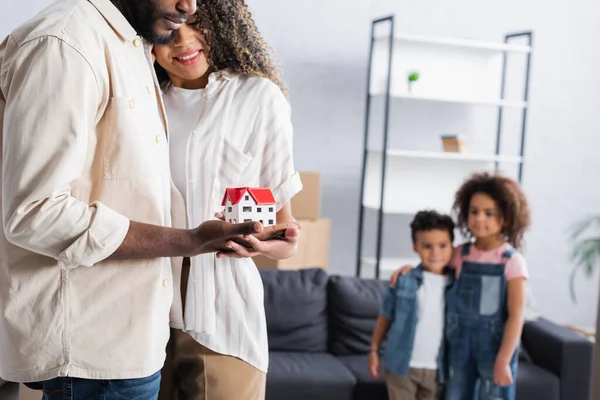 Smiling african american couple holding house model near blurred children in new apartment — Stock Photo