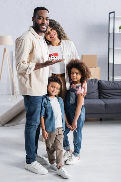 Feliz homem americano africano segurando casa modelo perto de esposa e filhos em novo apartamento — Fotografia de Stock