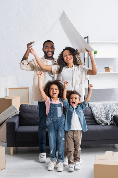 Crianças americanas africanas animado mostrando gesto de sucesso perto dos pais com telhado de papelão no novo apartamento — Fotografia de Stock