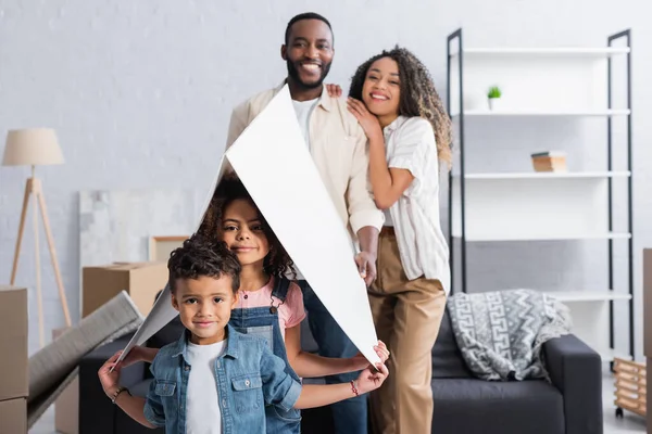 Bllured casal afro-americano sorrindo perto de crianças sob telhado de papel no novo apartamento — Fotografia de Stock