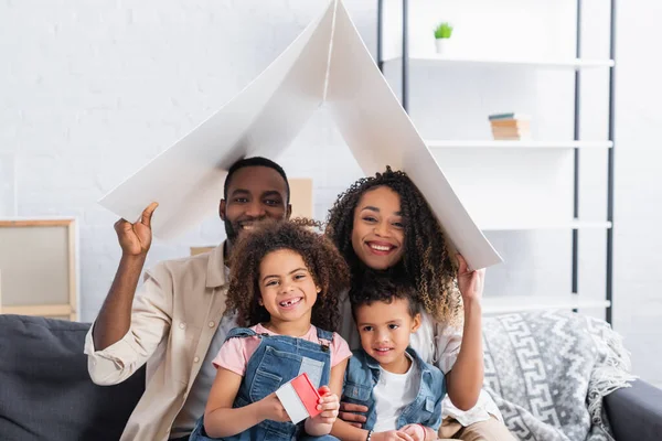 Familia afroamericana feliz mirando la cámara bajo techo de papel en nuevo apartamento - foto de stock