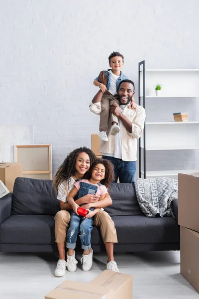 Alegre afroamericano hombre sosteniendo hijo en hombro cerca esposa e hija sentado en sofá - foto de stock