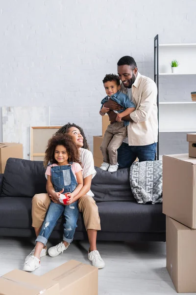 Afrikanisch-amerikanischer Mann mit Sohn schaut glückliche Frau an, die mit Tochter auf Sofa sitzt — Stockfoto