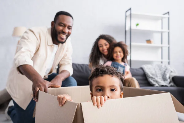 Africano americano ragazzo seduto in scatola di cartone vicino allegra famiglia su sfondo sfocato — Foto stock