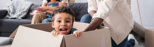 Thrilled african american boy sitting in carton box near blurred family, banner — Stock Photo