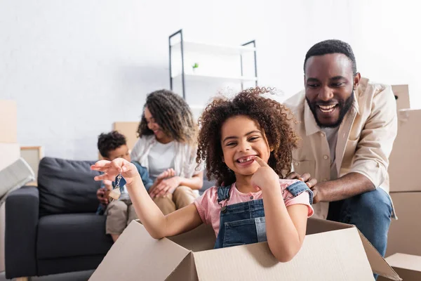 Chica afroamericana en caja de cartón sosteniendo la llave de nuevo apartamento cerca de papá feliz - foto de stock