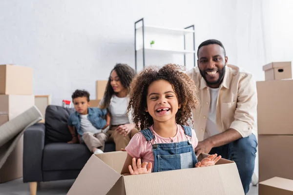 Eccitato africano americano ragazza ridendo in scatola di cartone vicino sfocato papà e mamma con fratello — Foto stock