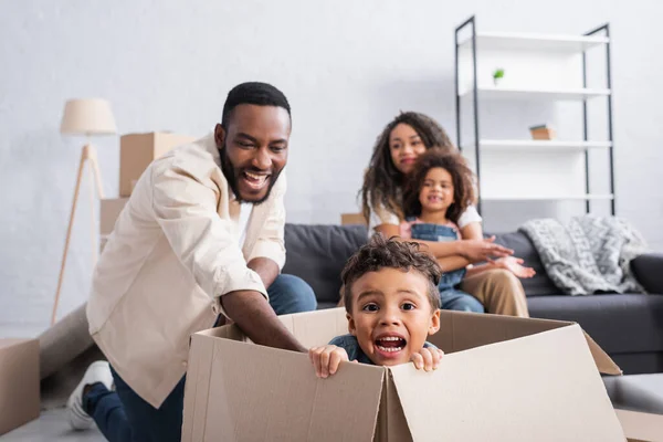 Garoto americano africano assustado na caixa de papelão perto da família alegre no novo apartamento — Fotografia de Stock
