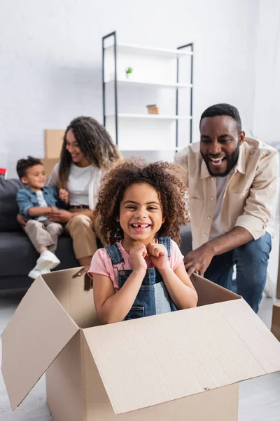 Felice ragazza africana americana in scatola di cartone vicino sorridente papà e mamma offuscata con fratello — Foto stock