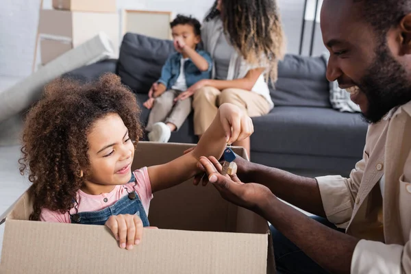 Afro-américaine fille dans la boîte en carton donnant la clé de papa heureux près de maman floue et frère — Photo de stock