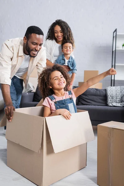 Весела афроамериканська дівчина розважається в картонній коробці біля щасливої сім'ї — стокове фото