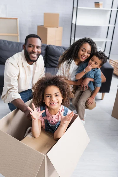 African american girl in carton box showing scaring gesture near blurred family in new apartment — Stock Photo