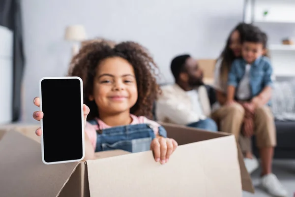 Gioiosa ragazza africana americana seduta in scatola di cartone e mostrando smartphone con schermo bianco — Foto stock