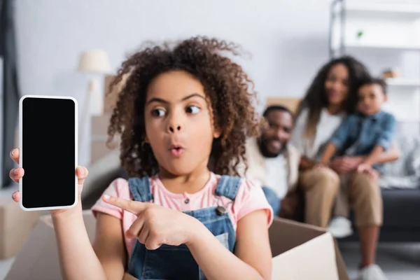 Enfant afro-américain étonné pointant vers le smartphone avec écran vide tout en étant assis dans une boîte en carton — Photo de stock