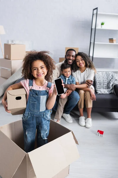 Bambino felice con scatola di cartone e cellulare con schermo bianco vicino alla famiglia afroamericana offuscata — Foto stock