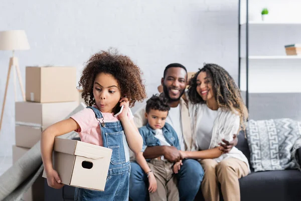 Fille surprise avec boîte en carton parlant sur téléphone mobile près de la famille afro-américaine floue — Photo de stock