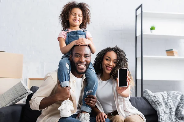 African american woman showing smartphone with blank screen near happy husband and daughter — Stock Photo